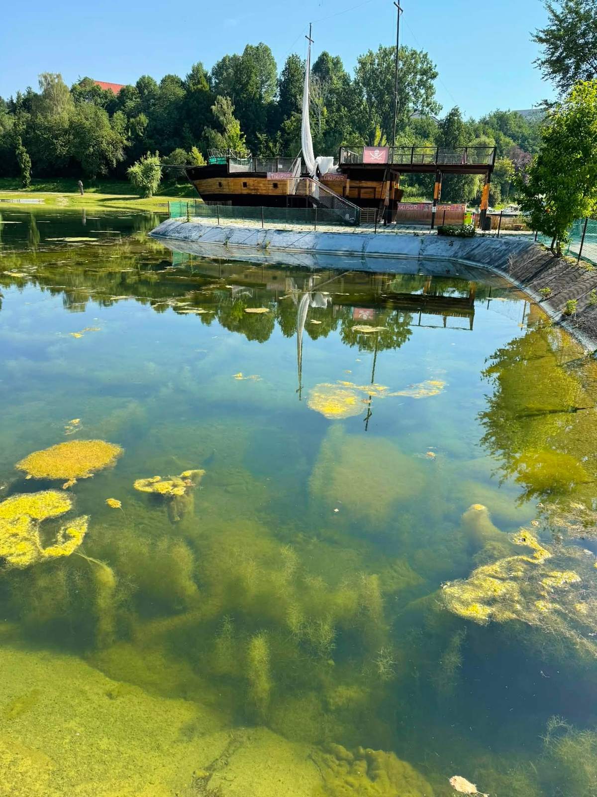Jazero so zelenými riasami na hladine, v pozadí drevená loď pri brehu