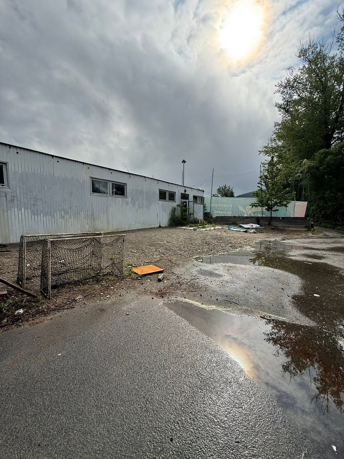 Nespevnená plocha s kalužami pred plechovou budovou, na zemi leží rozobraná bránka