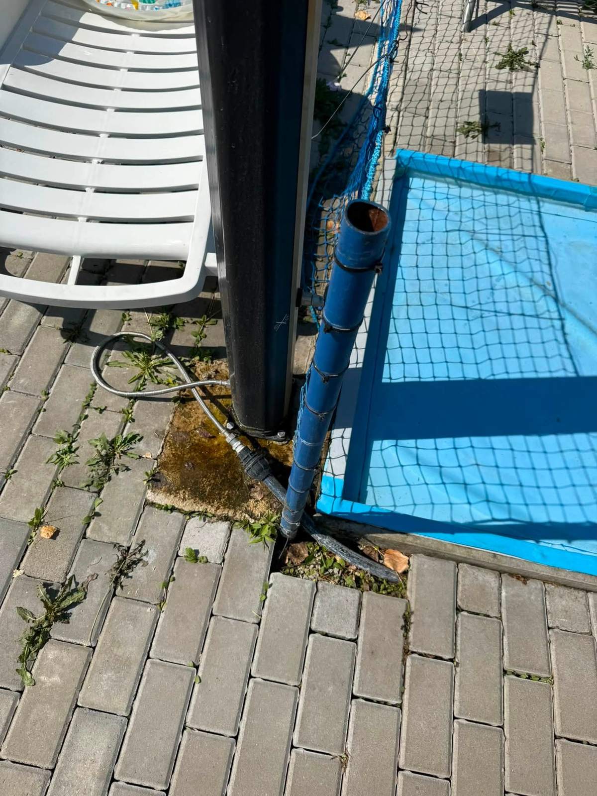 Kovový stĺp s ochrannou sieťou vedľa bazéna, po dlažbe vedie hadica, v škárach rastie burina